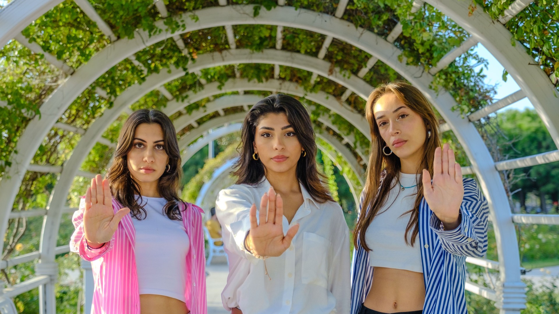 Three young female friends in a tree-filled park on a spring day, looking at the camera while making a "stop" gesture with their hands